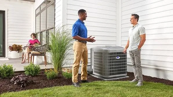 Technician in a blue uniform discussing an outdoor air conditioner with a homeowner beside the unit