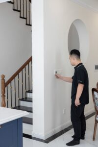 Man adjusting a wall thermostat in a hallway beside a staircase inside a house