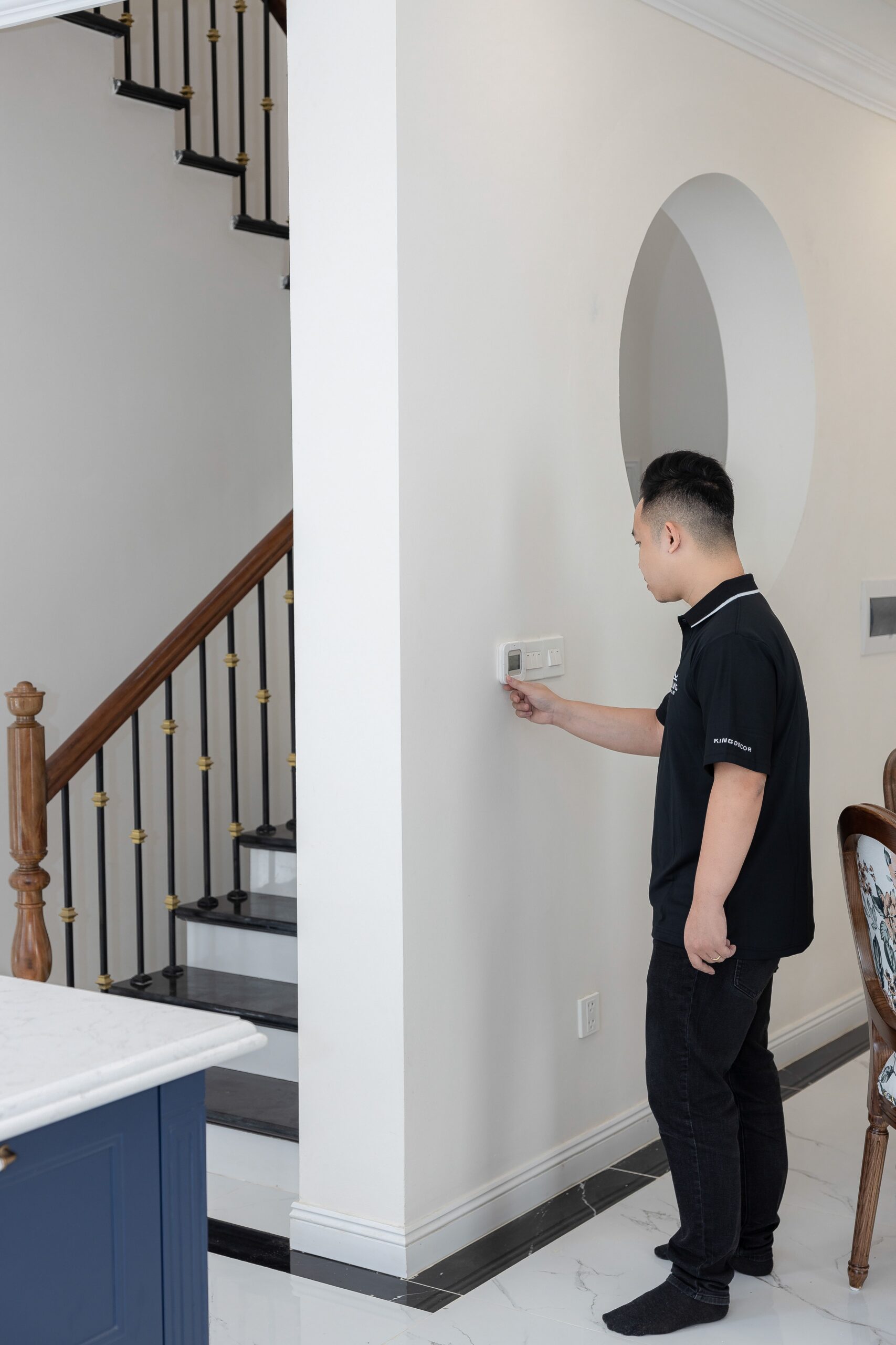 Man adjusting a wall thermostat in a hallway beside a staircase inside a house
