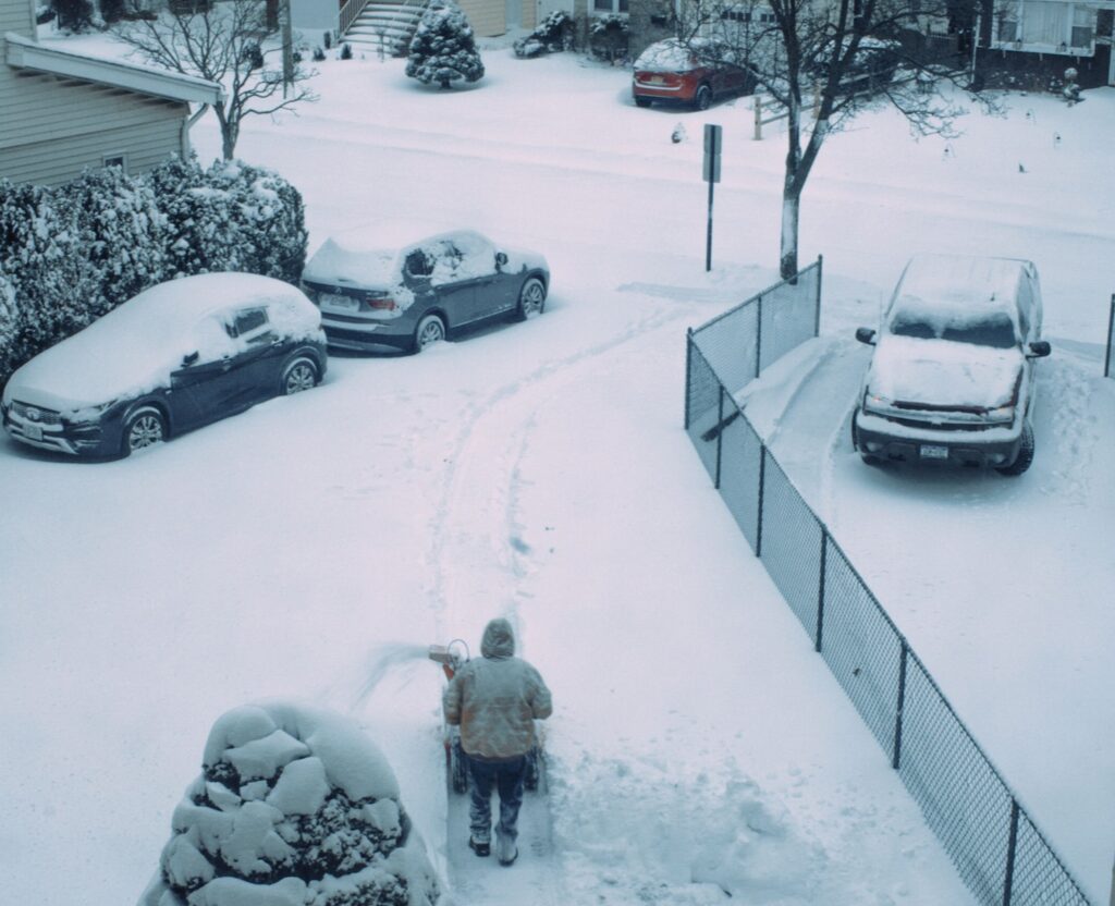Person shovelling a suburban driveway after heavy snow, with snow-covered cars along the street