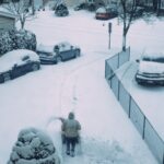 Person shovelling a suburban driveway after heavy snow, with snow-covered cars along the street