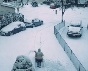 Person shovelling a suburban driveway after heavy snow, with snow-covered cars along the street